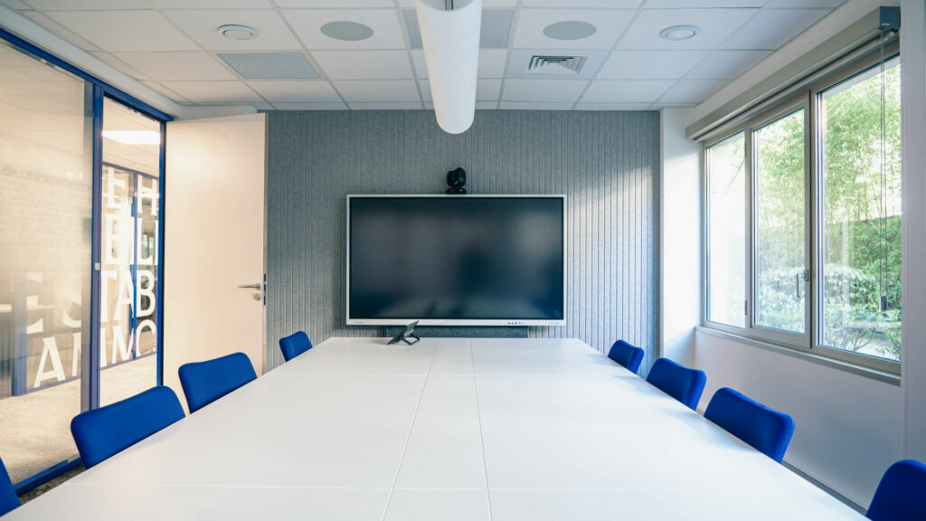 Grande salle de réunion connectée et moderne avec une longue table blanche, des chaises bleues, un grand écran interactif au mur pour la visioconférence et de grandes fenêtres donnant sur de la verdure.