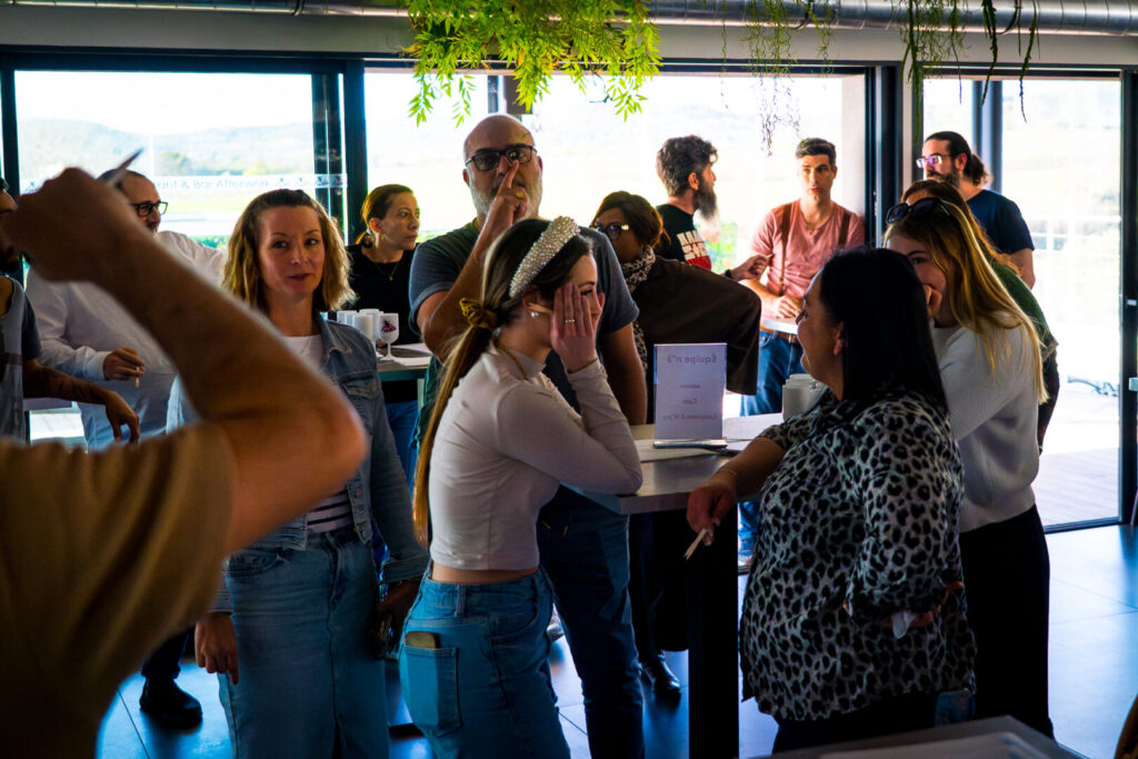 C'est une photo prise lors d'un atelier collaboratif en entreprise. Plusieurs collègues sont debout dans un espace de détente ou une salle de réunion aménagée, en train de participer à un défi de construction créatif. L'atmosphère est détendue et studieuse : chaque petit groupe est absorbé par la réalisation de sa structure en spaghettis. On sent une réelle émulation collective autour de ces tables hautes, avec des échanges et une cohésion visibles entre les participants. Le cadre est contemporain, avec une décoration soignée intégrant des éléments de verdure suspendus qui renforcent le côté chaleureux et accueillant du lieu.